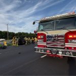 A fire truck with an American flag grill is parked on a highway near firefighters attending to a black car involved in an accident. Debris is scattered on the road under a blue sky with light clouds.