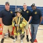 Four people pose indoors: three adults in black shirts, shorts or jeans, and hats, standing beside a smiling child who is wearing oversized firefighter gear and helmet. They are on a gym floor with blue bleachers behind them.