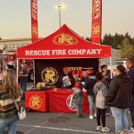 People gather around a red "Rescue Fire Company" tent at an outdoor event. Volunteers hand out items, and several children wear plastic fire helmets. Fire trucks and banners are visible in the background.