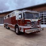 A large red and white fire rescue truck is parked on a concrete driveway in front of a brick building with several garage doors. The truck has “Cambridge” and an American flag on the front grille.