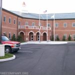 A brick building with arched entryways and a cupola, two flagpoles with flags, and a red fire chief SUV parked in front. The parking lot is mostly empty.