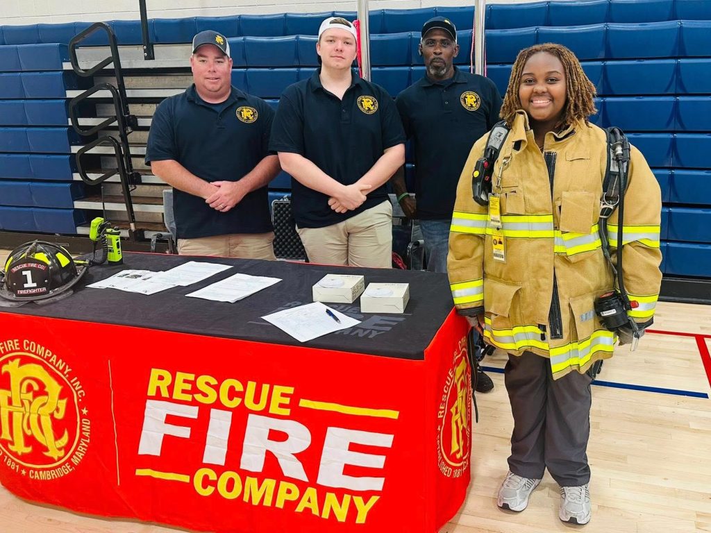 Four people stand behind a table with a red “Rescue Fire Company” banner; three wear matching shirts and one wears firefighter gear. Items, papers, and a helmet are on the table. Blue bleachers are in the background.